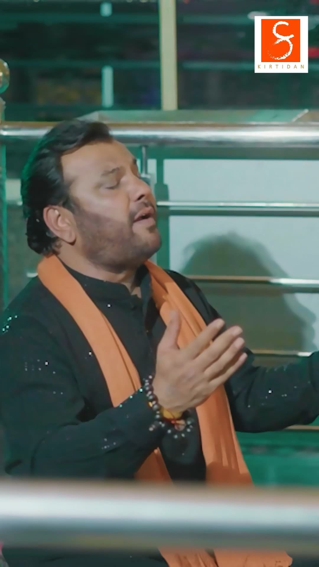 Kirtidan Gadhvi singing with hands raised, in a dark kurta and saffron scarf